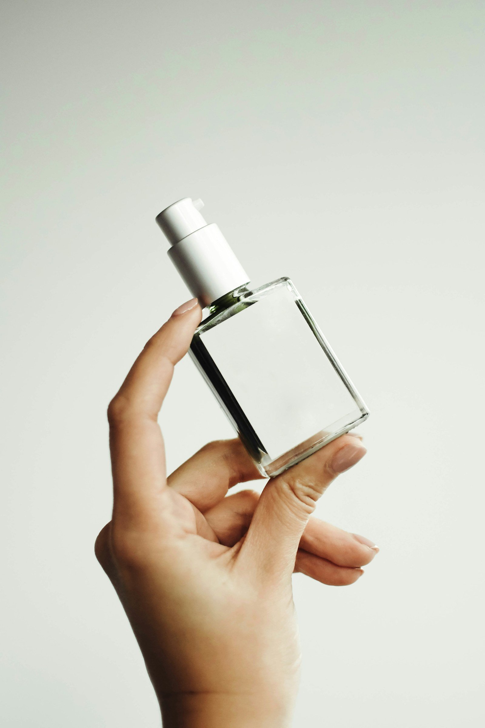 Tangan memegang botol parfum kaca bening tanpa label di depan latar terang