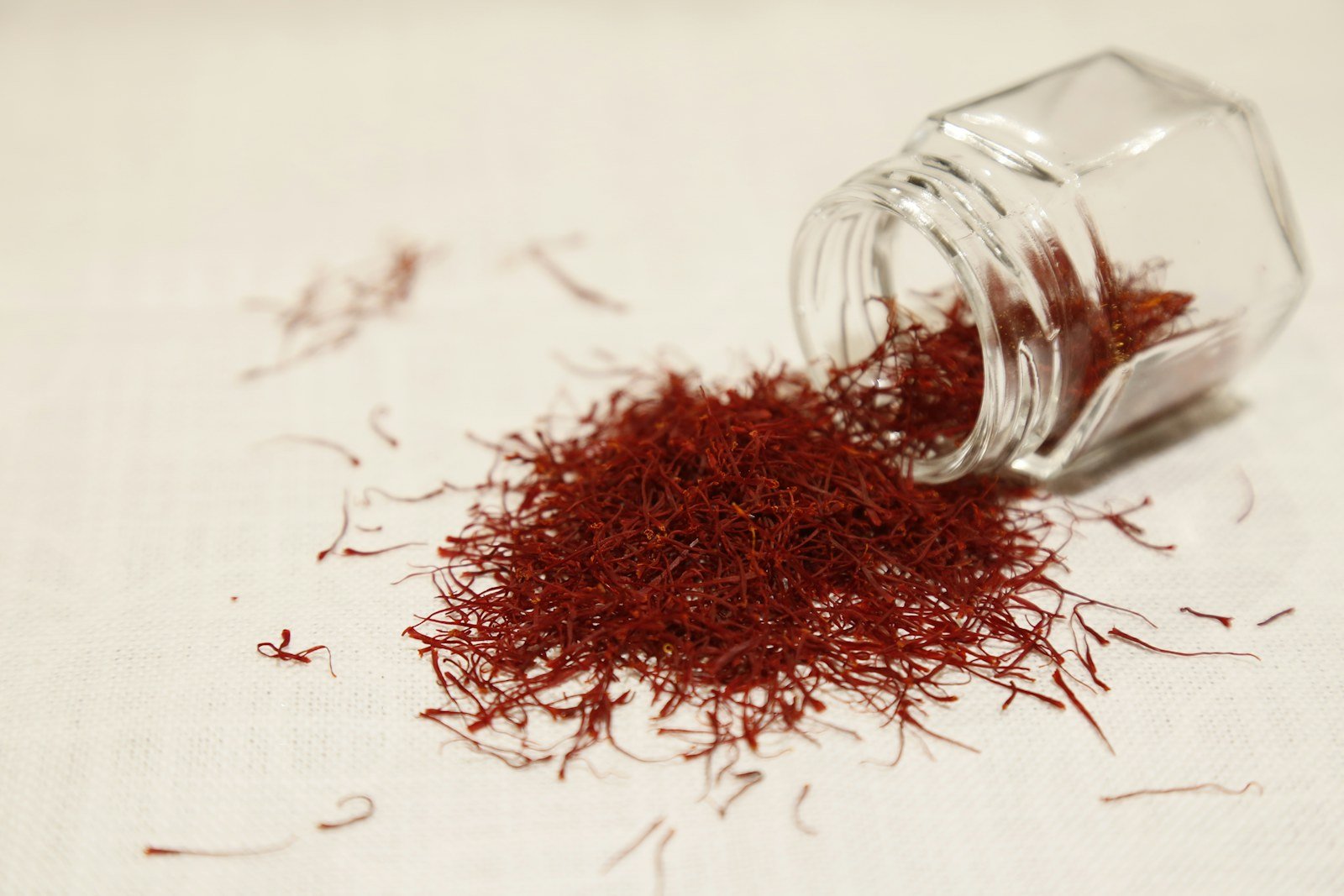 Helai saffron merah tumpah dari stoples kaca kecil di atas kain putih