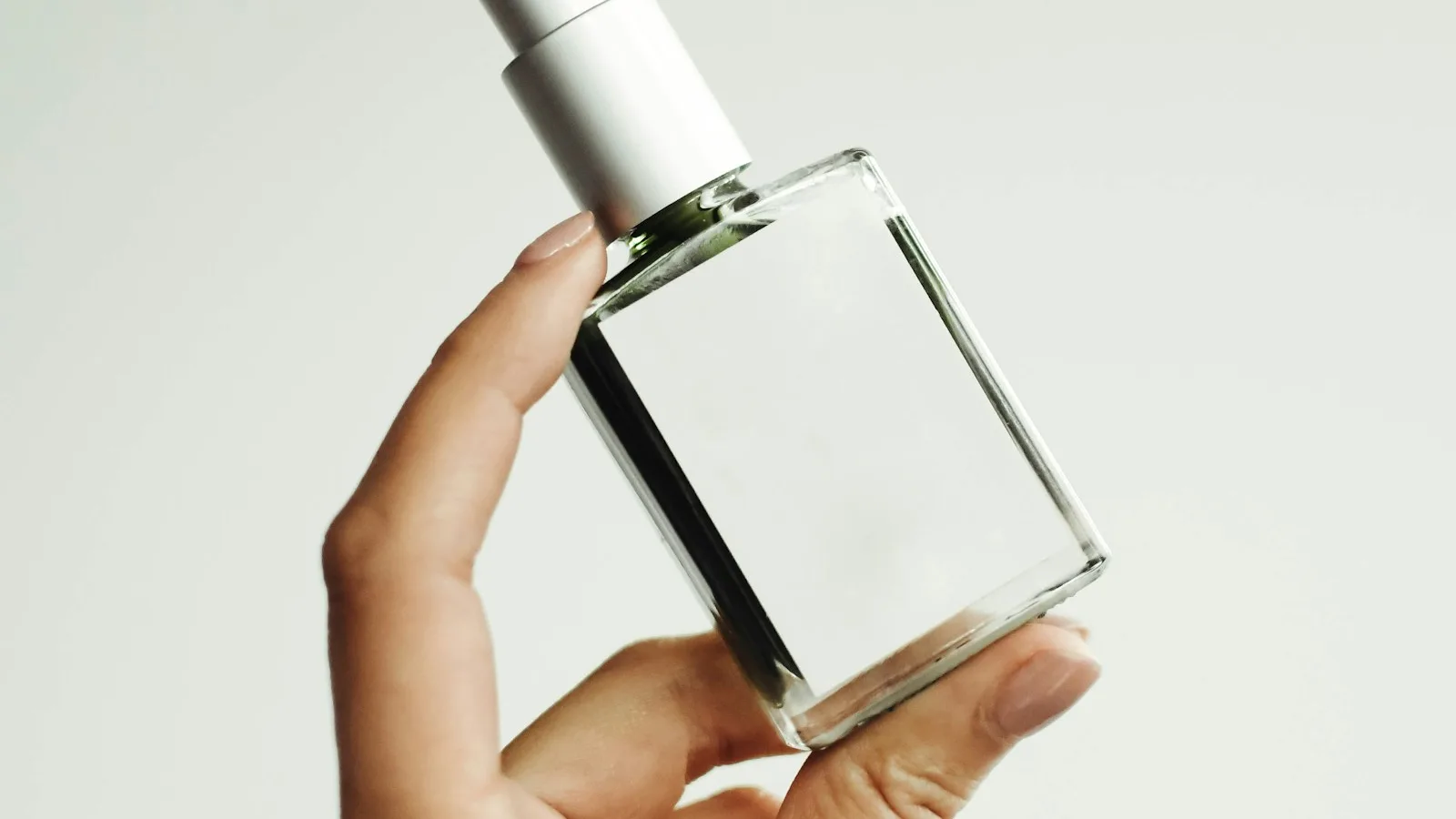 Tangan memegang botol parfum kaca bening tanpa label di depan latar terang