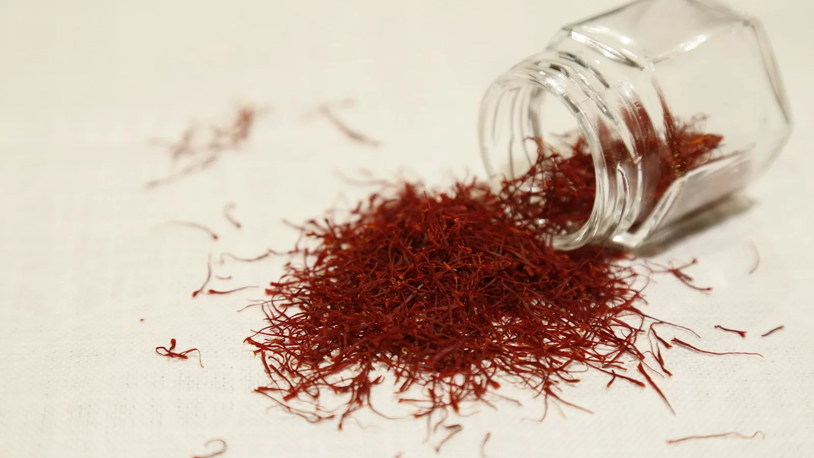 Helai saffron merah tumpah dari stoples kaca kecil di atas kain putih